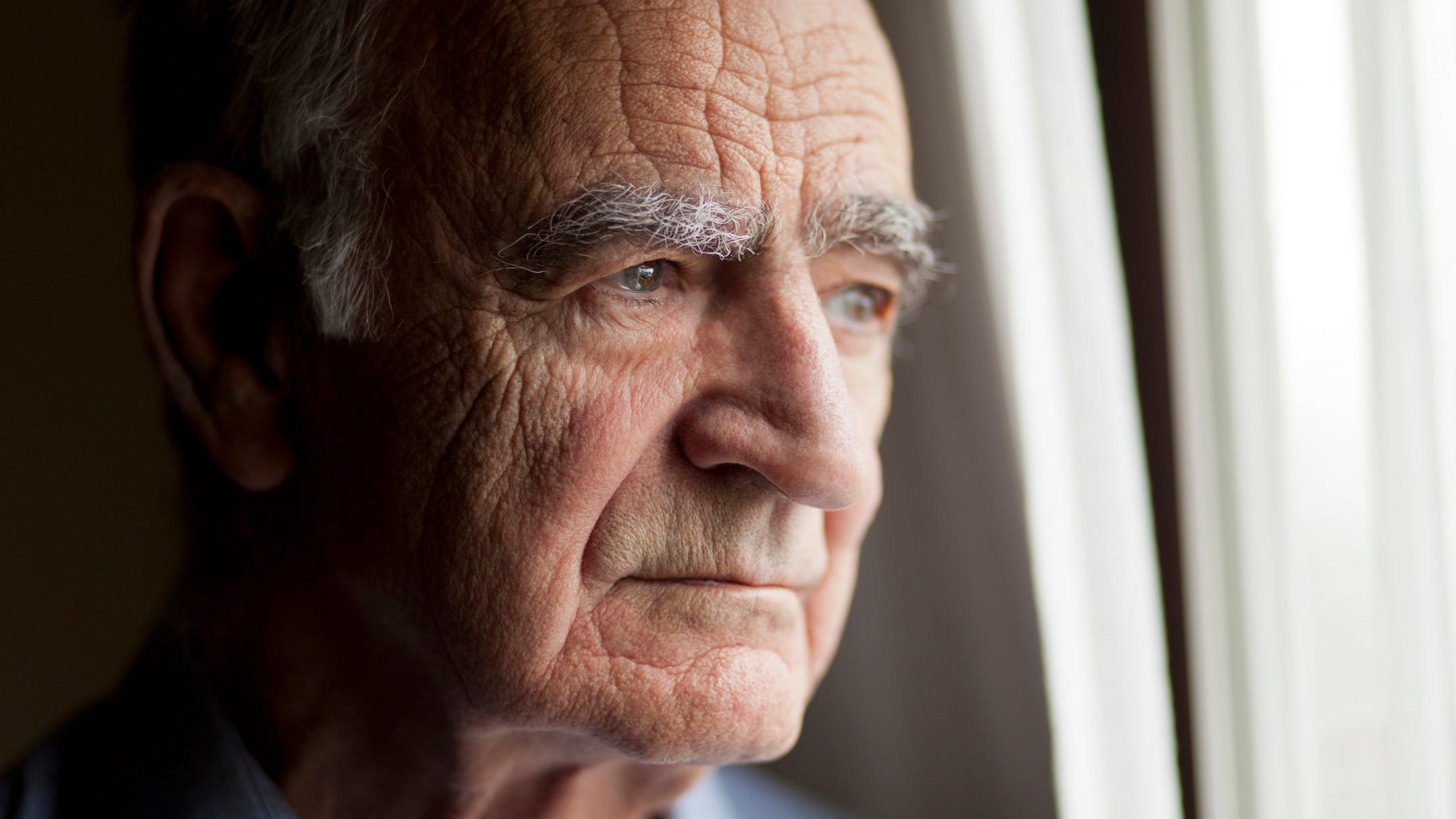 A visibly concerned senior citizen looking out a window.