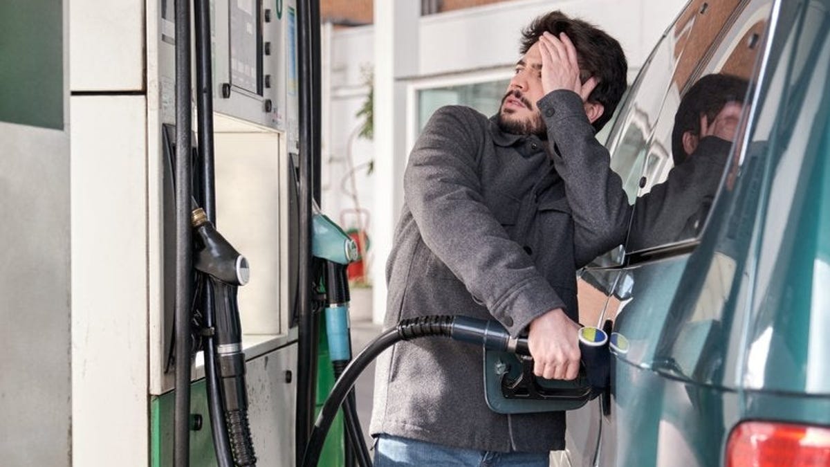 A young adult refuels their car while looking worried at the high gas prices at a gas station.