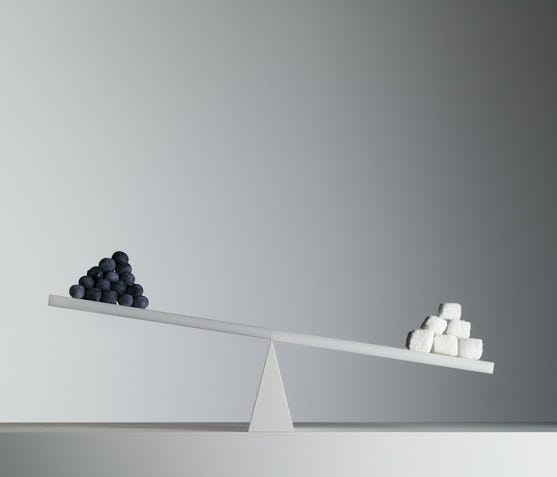 A seesaw with a pyramid of small black spheres on the left and larger white cubes on the right, against a blank gray wall.