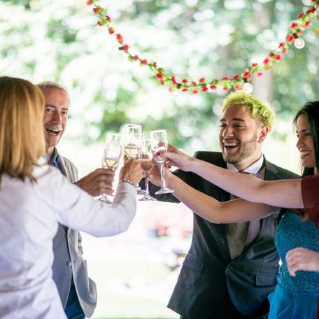 A group of wedding attendees in formal attire smile and toast their drinks.