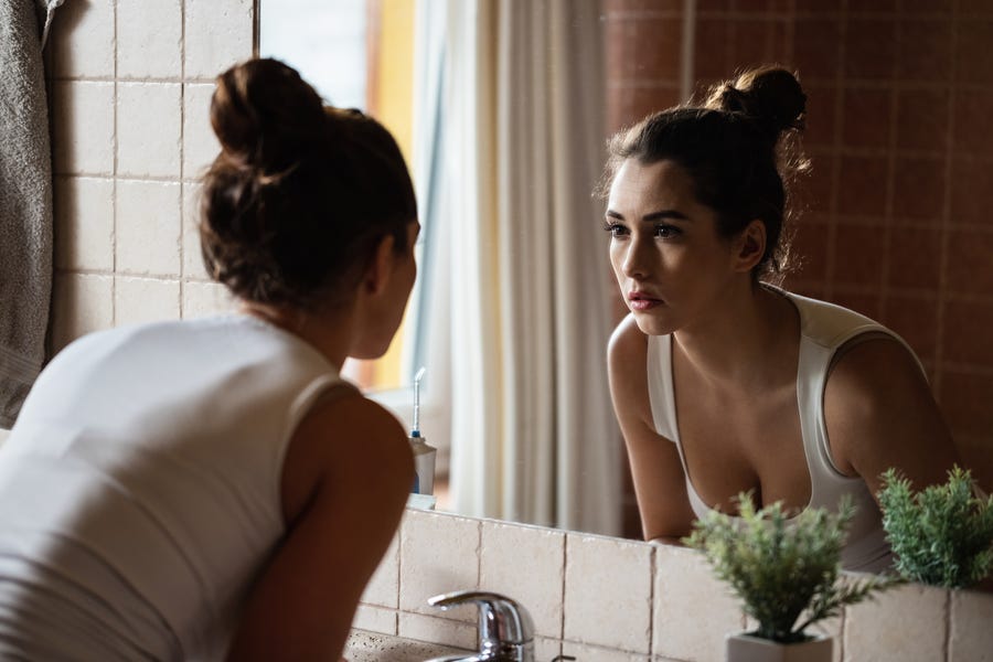 Pensive woman looking herself in the mirror in bathroom.