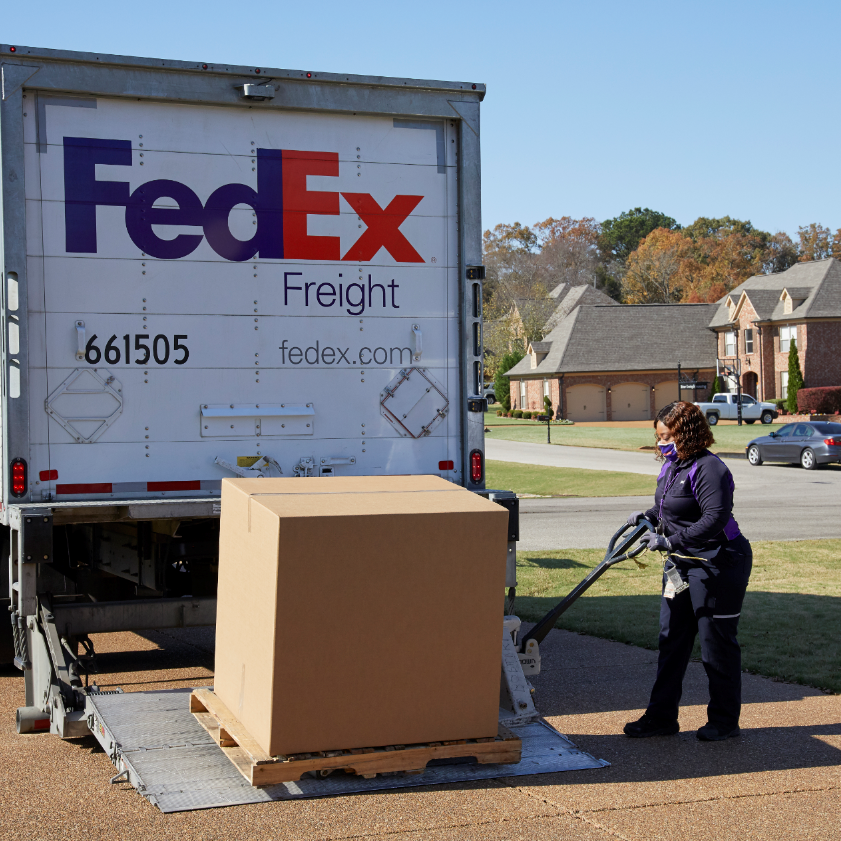 A FedEx Freight Direct employee unloads a large package onto a driveway in a residential neighborhood.