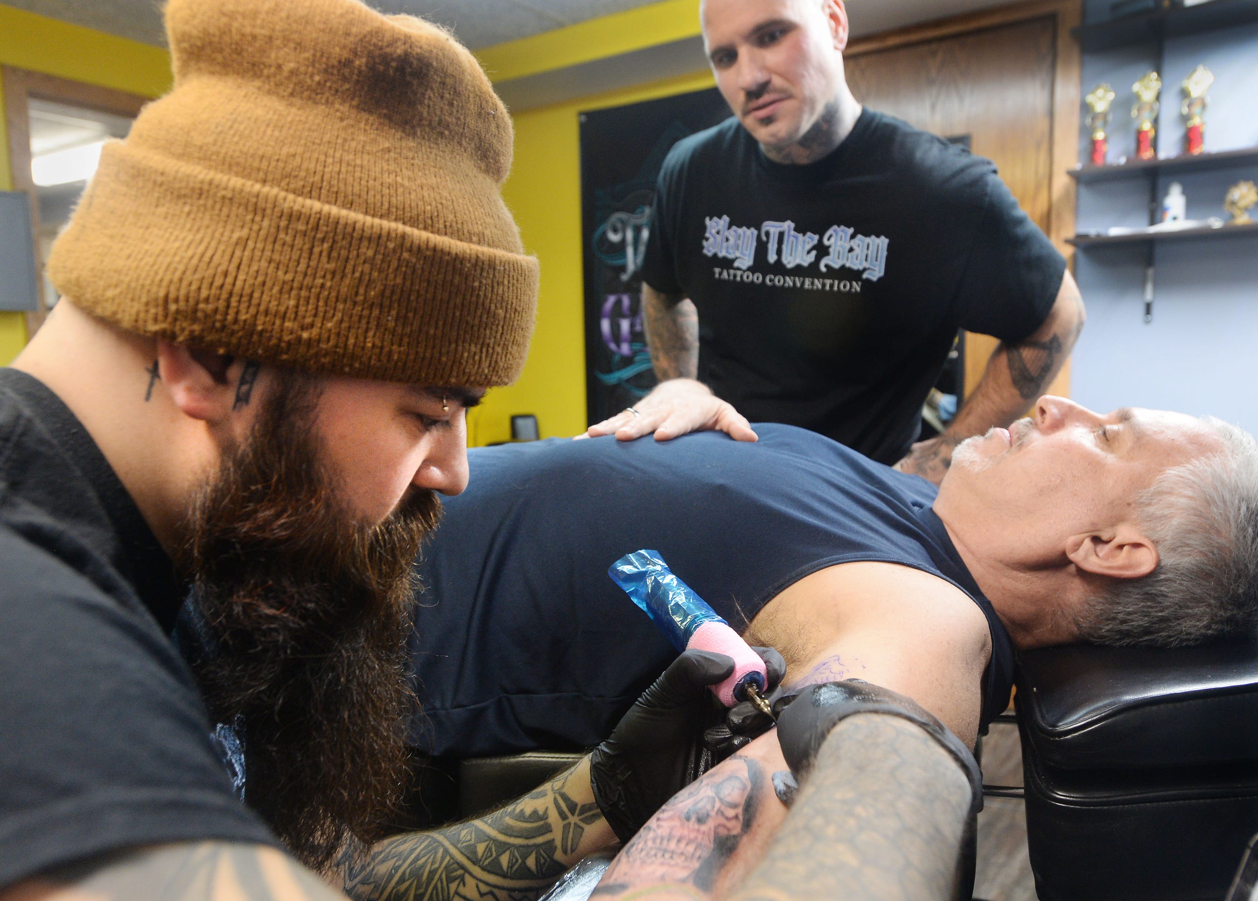 Tattoo artist Garrett Ybanez, left, works on a design for customer Tom Kolb, 59, right, on Dec. 14, 2021, at InkXpression tattoo shop in Millcreek Township. Ybanez, 30, and colleague Ryan Quinn, 32, center, are coordinating the upcoming "Slay the Bay" tattooing convention.